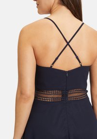 Sötétkék ruházat, keresztbe tett vékony pántokkal és textúrált csipke panellel a deréknál, rejtett cipzárral a hátsó részen.