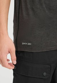 Camisa de secado rápido de color gris oscuro con mangas cortas, que presenta un detalle de logo en la costura lateral. La camisa se combina con pantalones cortos tipo cargo negros con bolsillos abotonados.