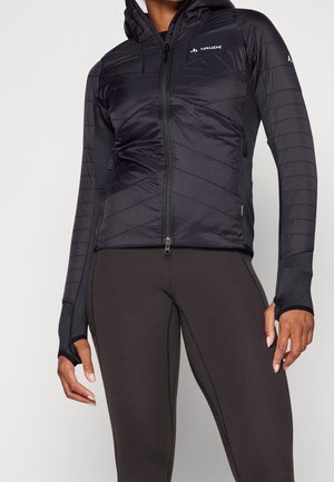 Femme portant une veste matelassée noire à capuche avec des trous pour les pouces et des leggings sombres ajustés, debout devant un fond uni.