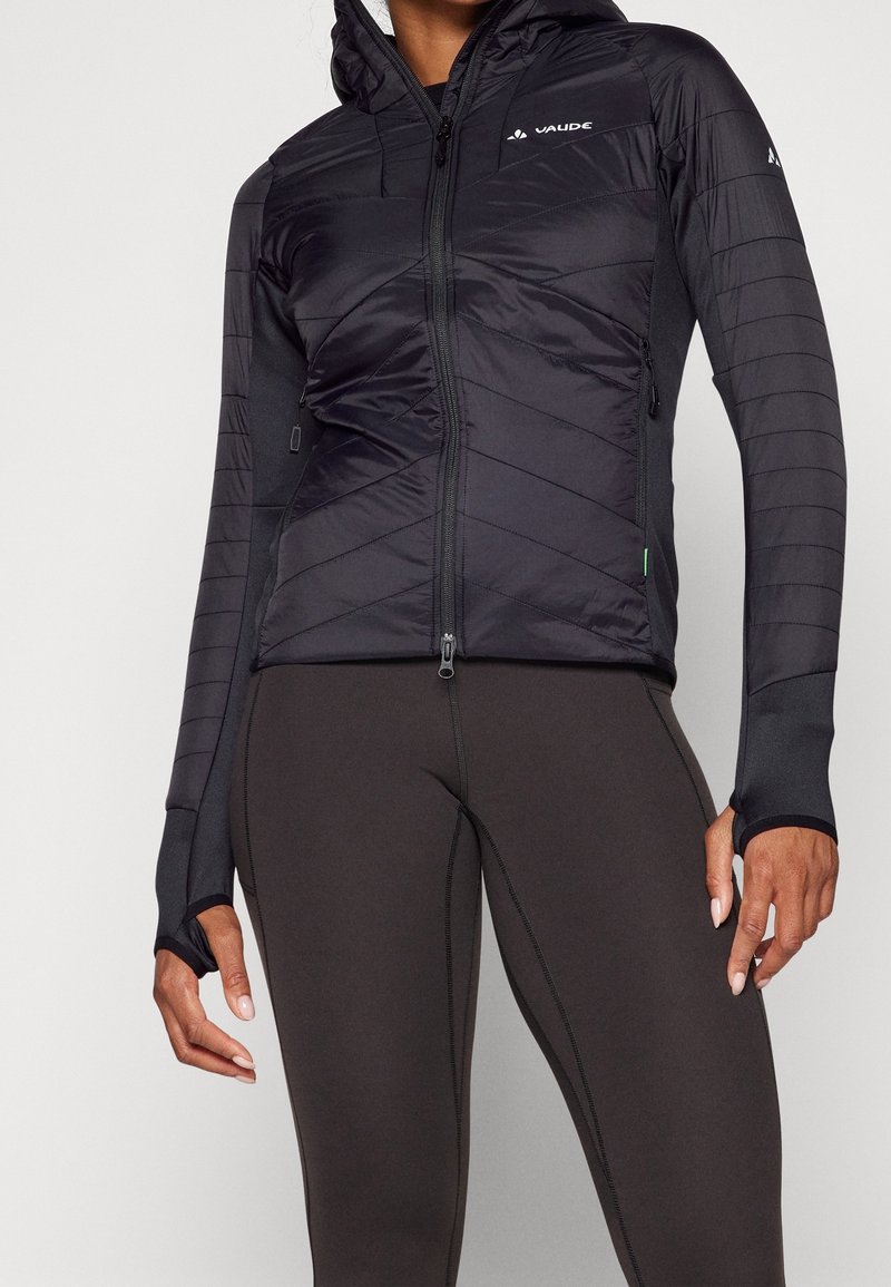 Femme portant une veste matelassée noire à capuche avec des trous pour les pouces et des leggings sombres ajustés, debout devant un fond uni.
