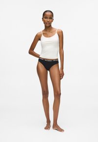 Kobieta stoi boso w białym podkoszulku i czarnych majtkach z markowym pasem na tle jednolitego białego tła.