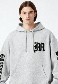 Felpa grigia con cappuccio e tasca frontale, caratterizzata da un grande logo nero a forma di "M" sul petto e grafiche testuali su entrambe le maniche.