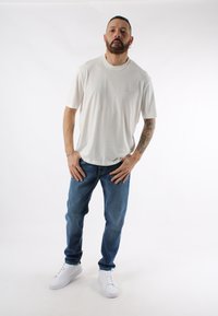 T-shirt a maniche corte, grigio chiaro, con un logo discreto, abbinato a jeans in denim blu e sneakers bianche. Taglio dritto, vestibilità casual, con texture minimale.