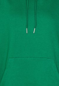 Sweat à capuche vert en tissu doux, avec une poche avant, des cordons de serrage réglables et des embouts argentés sur les cordons.