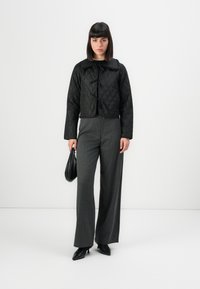 Femme portant une veste matelassée noire avec des liens, un pantalon gris à larges pattes, des talons noirs, tenant un petit sac à main noir, se tenant devant un fond blanc uni.