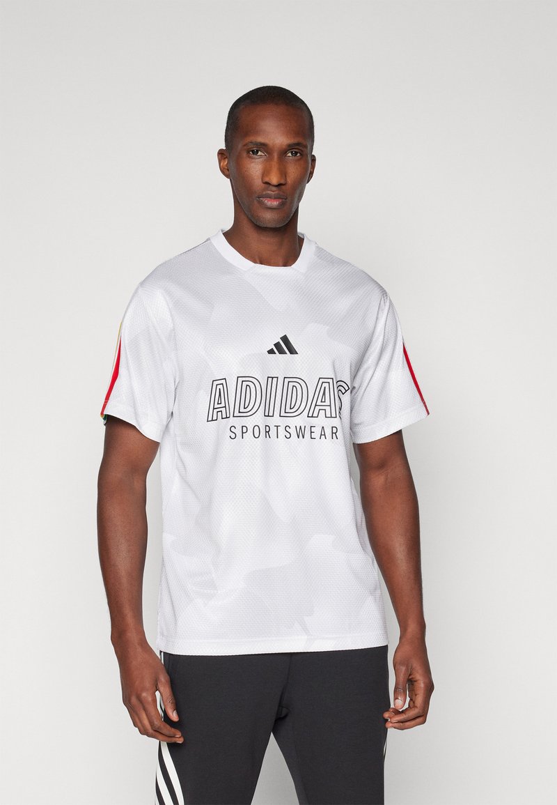 Balta acu sieta sporta T-kreklā ar melnu "ADIDAS SPORTSWEAR" logo, sarkaniem akcentiem uz piedurknēm un apaļu kakla izgriezumu. Viegls un elpojošs audums.