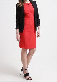 Rode nauwsluitende jurk met een uitsnijding bij de hals, gecombineerd met een zwarte blazer. Het model draagt zwarte sandalen met hoge hakken. Soepele stof, op maat gemaakt ontwerp.