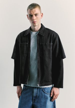 Jeune homme aux cheveux courts portant une veste noire sur une chemise grise et un jean bleu sur un fond clair uni.
