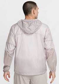 L�ätt, huva jacka i mjukt, ljusgrått material. Har elastiska manschetter och en rak fåll, designad med en slät, matt textur.