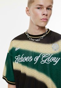 Groene, beige en zwarte grafische t-shirt met hexagonaal patroon en de tekst "Echoes of Glory", geribbelde kraag en zilveren ketting.
