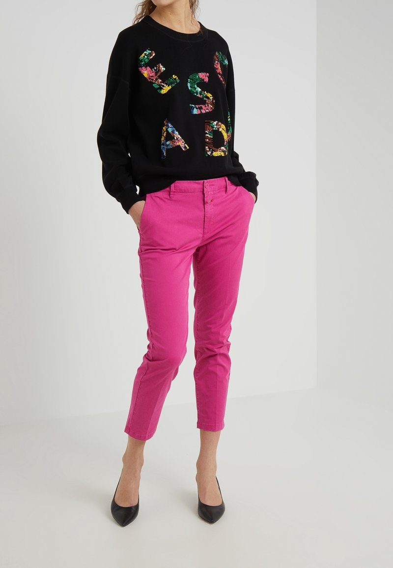Woman in bright pink cropped pants, black sequined sweatshirt with colorful letters, and black pointed-toe heels, standing with hands in pockets.