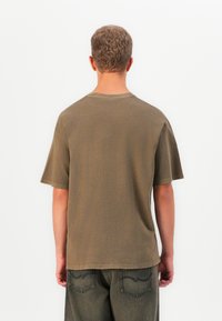 Mann med kort, krøllete hår iført en løs brun T-skjorte og falmede mørke jeans, stående mot en enkel hvit bakgrunn.