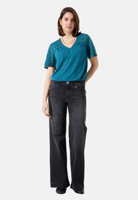 Teal kurzärmliges Top mit Spitzenverzierungen und V-Ausschnitt, kombiniert mit schwarzen weit geschnittenen Jeans. Glatte Textur, lässige Passform, keine Muster.