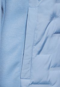 Světle modrá bunda s hladkou, polstrovanou strukturou a viditelným švem. Boční kapsa je integrována do designu, což zvyšuje funkčnost.