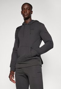 Felpa grigia scura con tasca frontale, cappuccio con cordini e logo ricamato. Il materiale è liscio, con pantaloni della tuta grigi abbinati visibili.