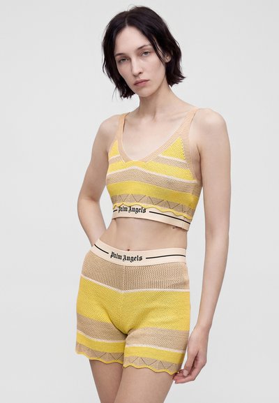 Γυναίκα που φοράει κίτρινο και μπεζ πλεκτό crop top και σετ σορτς με "Palm Angels" τυπωμένο στις ελαστικές ζώνες της μέσης, στέκεται μπροστά σε απλό φόντο.
