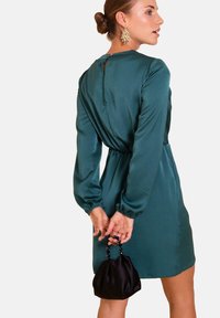 Abito verde in satin con maniche lunghe, vita cinchata e dettaglio a occhio di bue sulla schiena. La modella tiene una piccola borsa nera con manici di perline.
