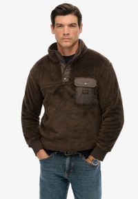Brun fleece pullover med kvart lynlås, med en struktureret overflade, en kontrasterende glat stofkappe og en frontlomme med trykknapper.