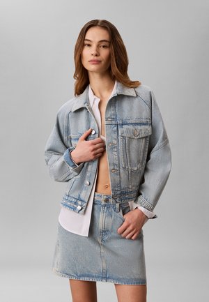 Frau mit hellbraunen Haaren trägt eine übergroße hellblaue Jeansjacke und passenden kurzen Jeansrock über einem teilweise aufgeknöpften weißen Hemd.