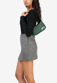 Sac à épaule synthétique vert en forme plate, avec un corps rectangulaire, accent logo et texture lisse, porté par une personne en haut noir et jupe à motifs.