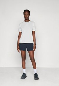 Helehall sportlik lühikeste varrukatega t-särk tumeroheliste lühikestega. Mustade tossude ja valgete sokkidega, seisab neutraalsel taustal.