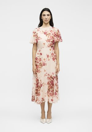 Γυναίκα με μακριά σκούρα μαλλιά στέκεται φορώντας ένα ανοιχτό ροζ μίντι φόρεμα με κόκκινο λουλουδένιο σχέδιο και φουσκωτά μανίκια, συνδυασμένο με λευκές μυτερές γόβες.