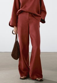 Pantalon large en lin couleur rouille assorti d'un haut oversize de la même teinte, accessoirisé d'un sac à main marron texturé avec des perles. Sandales plates noires portées.