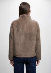 Brown faux-fur jacket with a collar, long sleeves, and soft texture. Worn with dark blue denim jeans, showing a casual style.