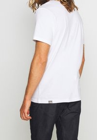 En person med långt hår och skägg, klädd i en enkel vit t-shirt med kort ärm och mörka byxor, som visas från baksidan och sidan.