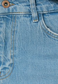 Telas de mezclilla azul claro con una superficie texturizada. Presenta costuras doradas y un botón de metal. Incluye un detalle de diseño de un bolsillo frontal.