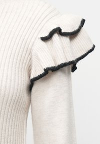 Svetlo bež pulover z rebrasto teksturo in naborkom na robovih rokavov. Naborki imajo kontrastno črno obrobo za dodatno podrobnost.