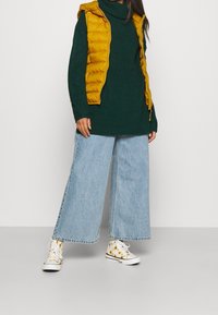 Maglione verde lavorato a maglia, gilet imbottito senape, jeans larghi blu e sneakers a fantasia gialla. Presenta un collo alto e una vestibilità rilassata.