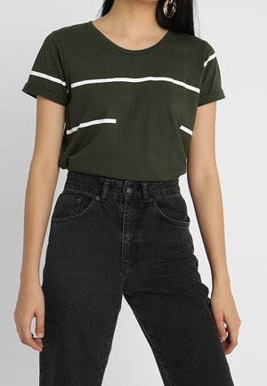 Kvinde iført en olivengrøn t-shirt med hvide striber, stukket ned i sorte højttaljede jeans, der viser talje og øvre ben.