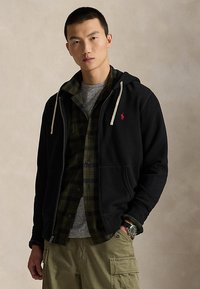 Jeune homme portant un sweat à capuche noir zippé par-dessus une chemise à carreaux verte et un t-shirt gris, avec un pantalon cargo kaki, debout devant un fond neutre.