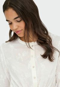 Blouse blanche brodée avec un motif floral texturé, encolure froncée et devant à boutons. Tissu en coton, léger et semi-transparent.