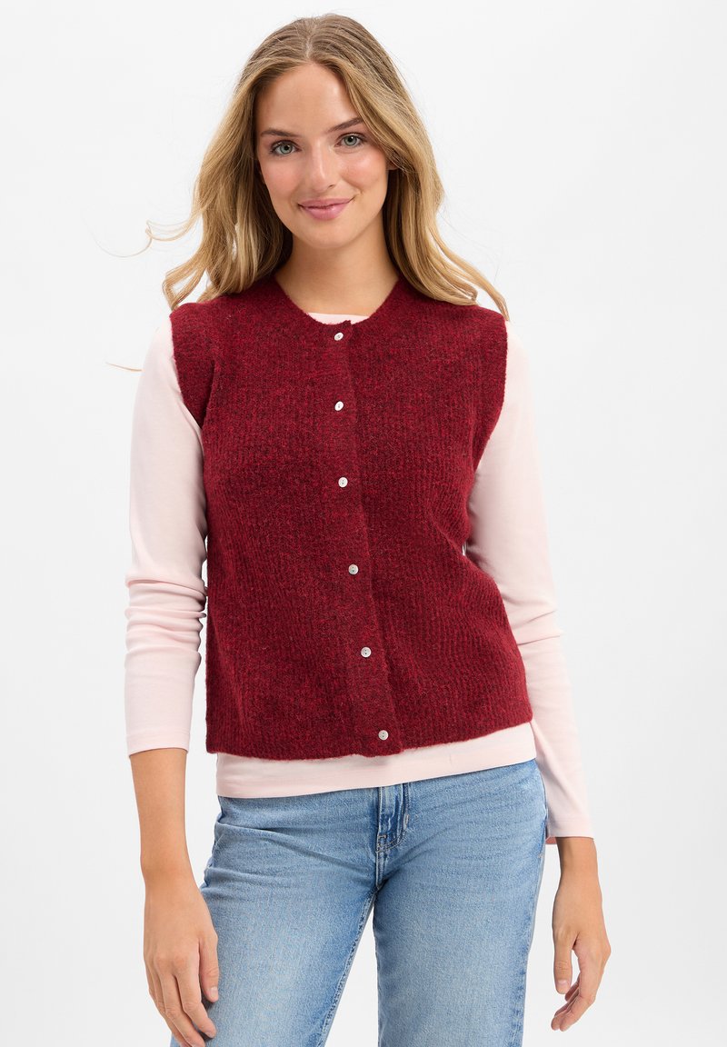 Czerwony sweter bez rękawów z okrągłym dekoltem i pięcioma pasującymi guzikami. Noszony na jasnoróżowej koszuli z długim rękawem. Faktura materiału wydaje się miękka.