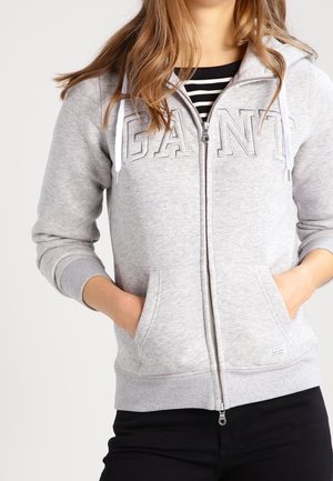 Woman wearing light grey zip-up hoodie with "GANT" embroidered on the chest, hands in pockets, and a black-and-white striped shirt underneath.