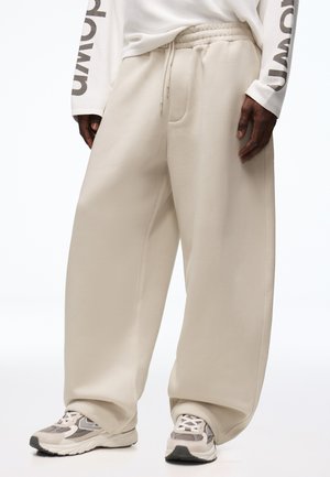 Persona che indossa pantaloni della tuta beige oversize con vita elastica e coulisse, abbinati a scarpe sportive bianche e grigie.