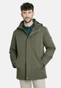 Hombre con cabello corto y rizado, vistiendo una chaqueta de invierno verde oliva sobre una camisa verde oscuro, con las manos en los bolsillos de la chaqueta, de pie contra un fondo blanco.