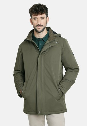 Hombre con cabello corto y rizado, vistiendo una chaqueta de invierno verde oliva sobre una camisa verde oscuro, con las manos en los bolsillos de la chaqueta, de pie contra un fondo blanco.