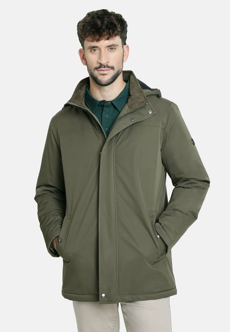 Hombre con cabello corto y rizado, vistiendo una chaqueta de invierno verde oliva sobre una camisa verde oscuro, con las manos en los bolsillos de la chaqueta, de pie contra un fondo blanco.