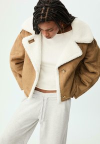 Persona con capelli intrecciati che indossa orecchini a cerchio, una giacca shearling beige, una maglietta bianca e pantaloni sportivi grigi chiari, che guarda verso il basso.