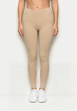 Leggings beige con un ajuste ceñido, fabricados con material elástico, que presentan una textura suave y una cintura alta, combinados con zapatillas blancas.