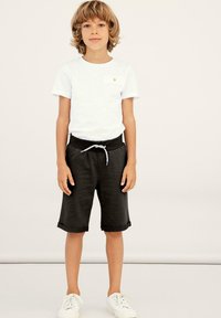 Child wearing a white short-sleeve T-shirt and black athletic shorts with a drawstring, paired with white sneakers.