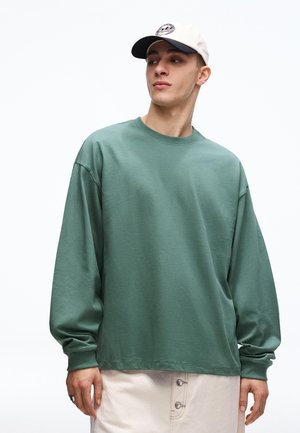 Mlajši moški, oblečen v dolgo majico zelene barve žajblja, bež hlače z gumbi in belo baseball kapo, ki gleda v stran na belem ozadju.