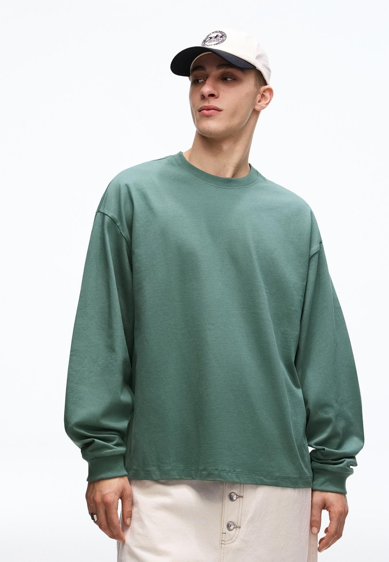 Mladý muž v šalviovo zelenej košeli s dlhým rukávom, béžových gombíkových nohaviciach a bielej šiltovke, ktorý sa pozerá stranou na bielom pozadí.