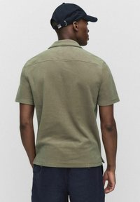 Lyhythihainen poolopaita oliivinvihreänä, pehmeästä kankaasta valmistettu. Siinä on klassinen kaulus ja suora helma, jossa on vähän yksityiskohtia.