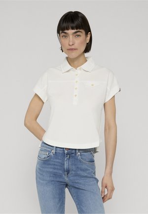 Frau mit kurzen dunklen Haaren, die ein weißes kurzärmeliges Poloshirt und blaue Jeans trägt, steht vor einem schlichten hellgrauen Hintergrund.