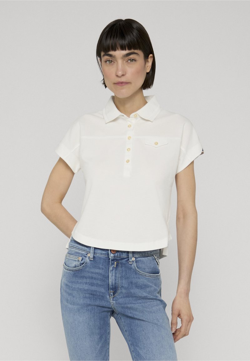 Femme aux cheveux courts et foncés portant un polo blanc à manches courtes et un jean bleu, debout devant un fond gris clair uni.