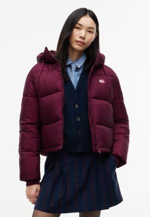 Femme portant une doudoune bordeaux, un cardigan bleu marine, une chemise bleu clair collet avec cravate, et une jupe plissée foncée sur fond blanc.